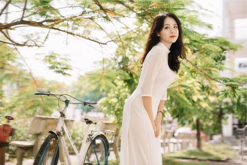 Hình ảnh bạn nữ mặc áo dài họa tiết hoa nhẹ, tạo dáng bên hàng cây trong sân trường.